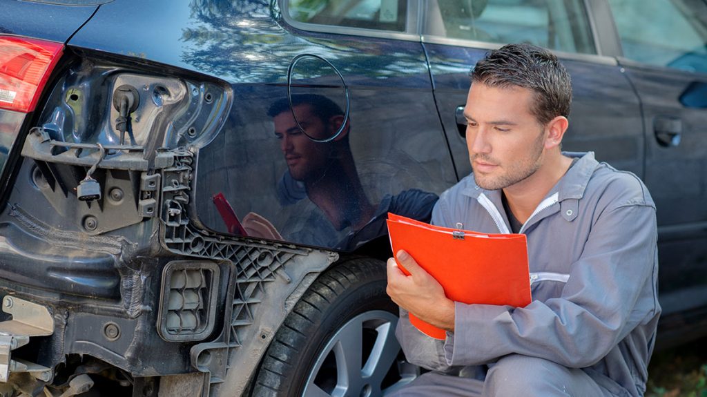 A collision repair employee taking notes on a clipboard next to a damaged vehicle with bumper removed